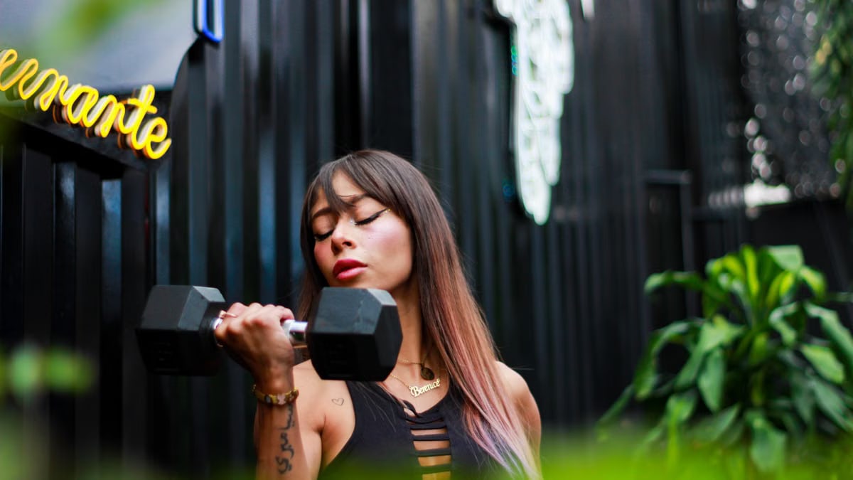 Fit woman with long hair and tattoos lifting a dumbbell with intense focus outdoors.